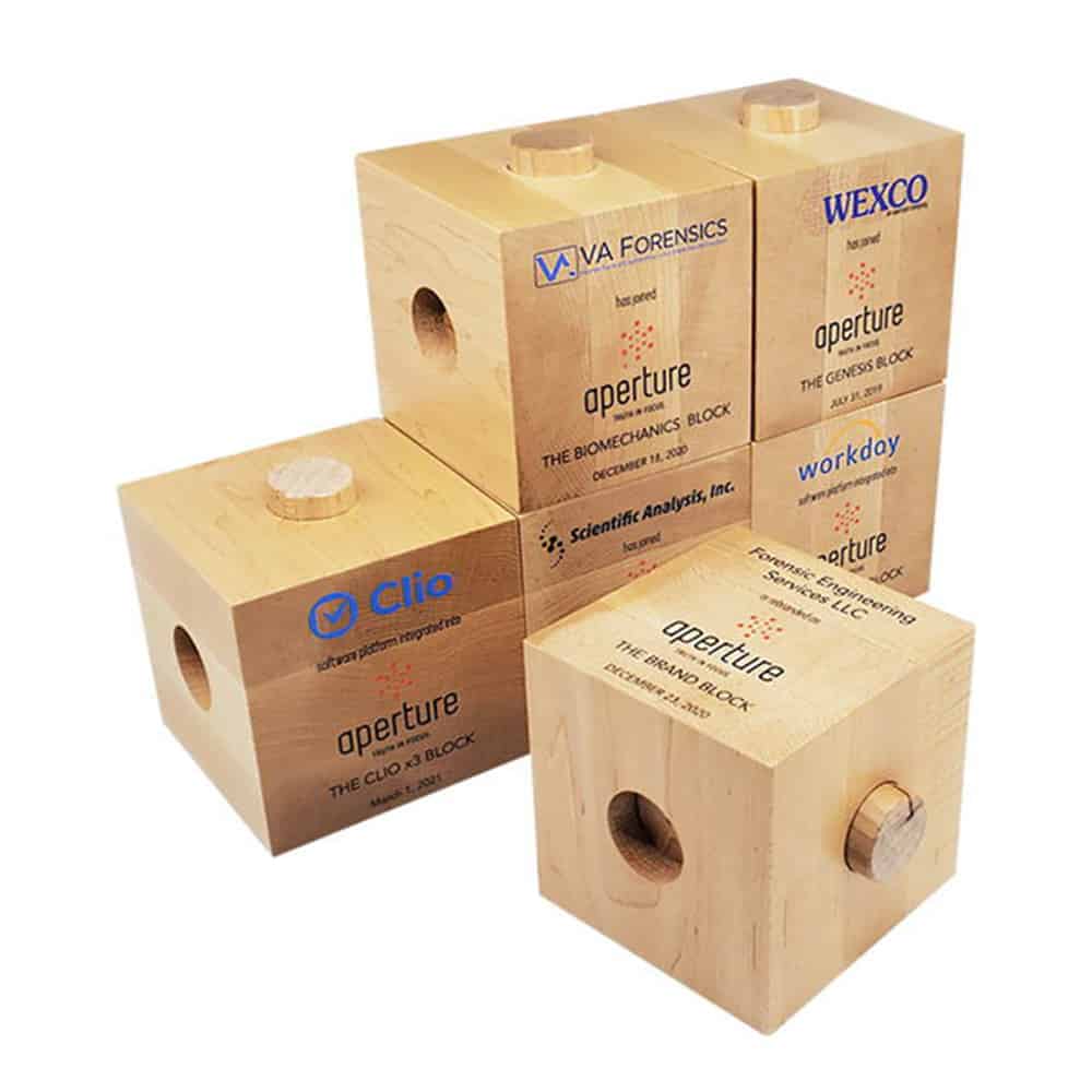 Wooden block deal toys featuring various company logos including Viva Forensics, Wexco, Clio, Workday, and Scientific Analysis, commemorating different partnerships and achievements with Aperture.